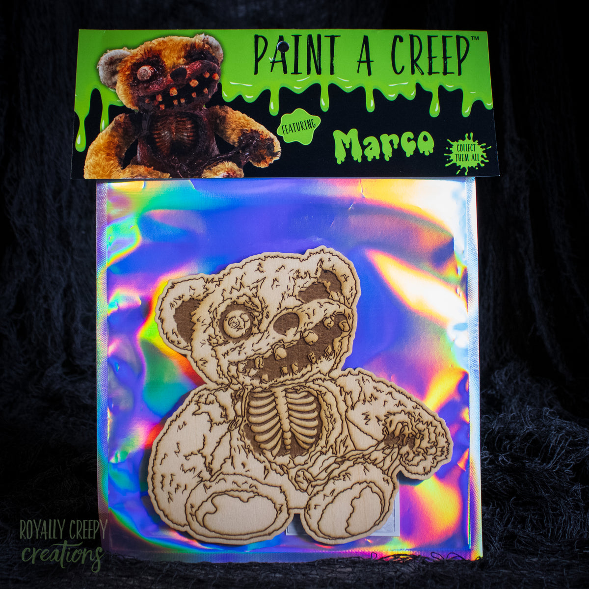 A packaged wooden bear craft kit with a laser-cut teddy bear design on an iridescent backing sheet, plus a branded header card reading 'Paint a Creep' and 'Marco'. The product is photographed against a dark textured background.