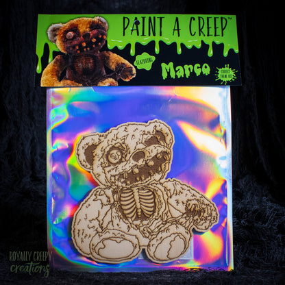 A packaged wooden bear craft kit with a laser-cut teddy bear design on an iridescent backing sheet, plus a branded header card reading 'Paint a Creep' and 'Marco'. The product is photographed against a dark textured background.
