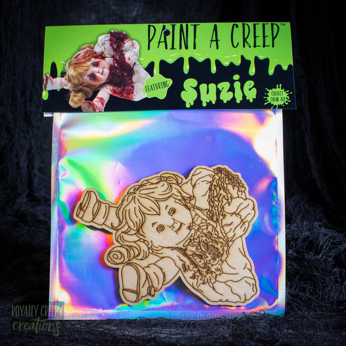 A wooden craft cutout of a creepy doll-like figure labeled 'Suzie' on an iridescent backing card, with a bright green header card reading 'PAINT A CREEP' and 'Suzie' at the top. The package is photographed against a dark textured fabric background.