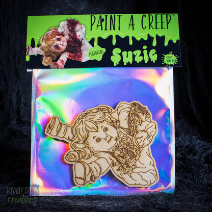 A wooden craft cutout of a creepy doll-like figure labeled 'Suzie' on an iridescent backing card, with a bright green header card reading 'PAINT A CREEP' and 'Suzie' at the top. The package is photographed against a dark textured fabric background.
