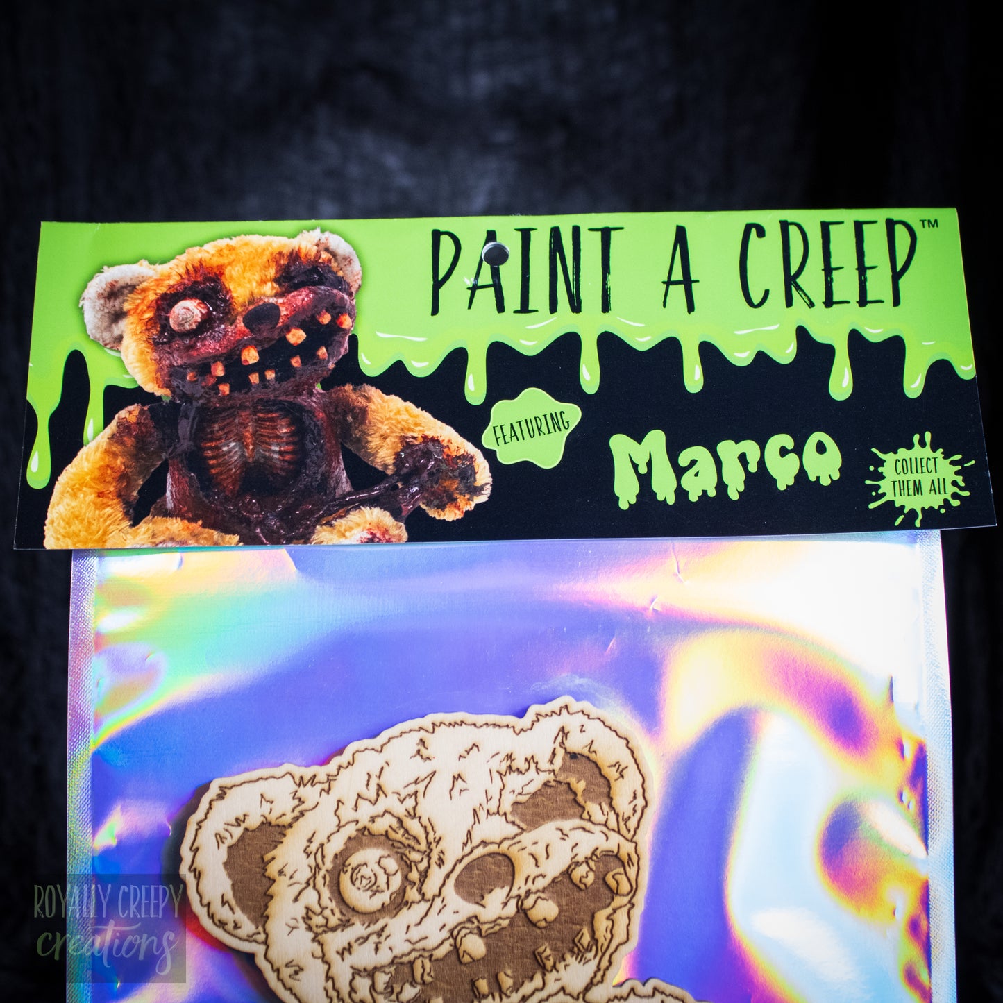 A packaged craft kit labeled 'PAINT A CREEP' featuring a creepy teddy bear design and the name 'Marco' on a bright green header card above an iridescent plastic bag containing a laser-cut bear face.