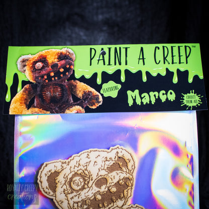 A packaged craft kit labeled 'PAINT A CREEP' featuring a creepy teddy bear design and the name 'Marco' on a bright green header card above an iridescent plastic bag containing a laser-cut bear face.
