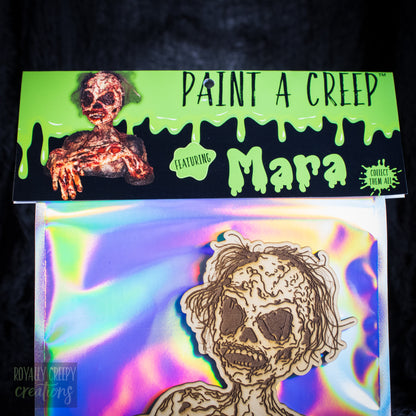 A packaged craft item showing a laser-cut wooden skeleton/creeper figure on an iridescent backing card. The top header reads 'PAINT A CREEP' and 'Mara'.