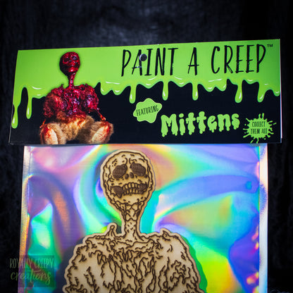 A packaged craft item showing a laser-cut wooden skeleton bearon an iridescent holographic backing card, with a branded header card reading 'Paint a Creep Mittens' against a black background.