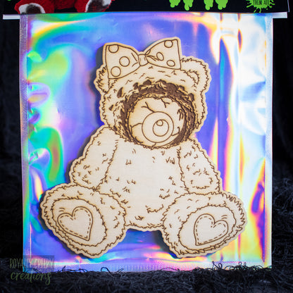 A laser-cut wooden teddy bear ornament with a polka-dot bow and heart-shaped feet, displayed on a colorful iridescent holographic backing sheet.