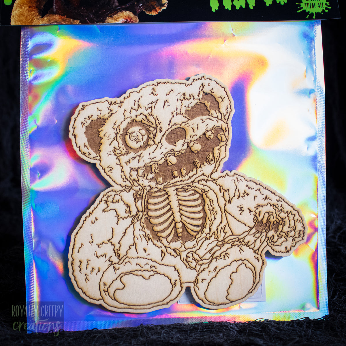A laser-cut wooden skeleton bear craft kit on an iridescent holographic backing sheet, photographed against a dark textured background.