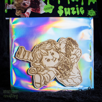 A wooden laser-cut doll design labeled 'Suzie' with a green slime-themed header card, displayed on an iridescent holographic backing over a dark textured surface.
