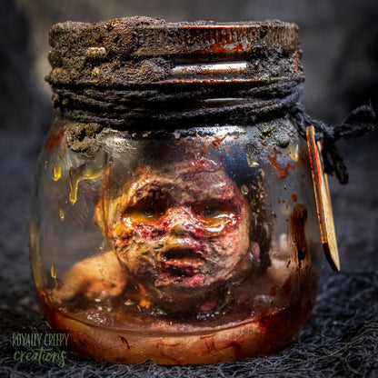 A creepy specimen prop of a severed doll head inside a dirty, rusted glass jar with a dark lid and rope, set against a dark textured background.