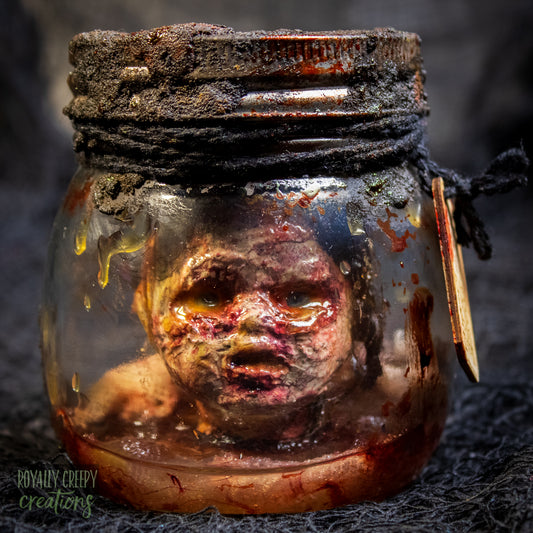 A creepy specimen prop of a severed doll head inside a dirty, rusted glass jar with a dark lid and rope, set against a dark textured background.