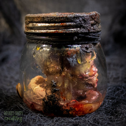 A creepy specimen prop of a severed doll head inside a dirty, rusted glass jar with a dark lid and rope, set against a dark textured background.