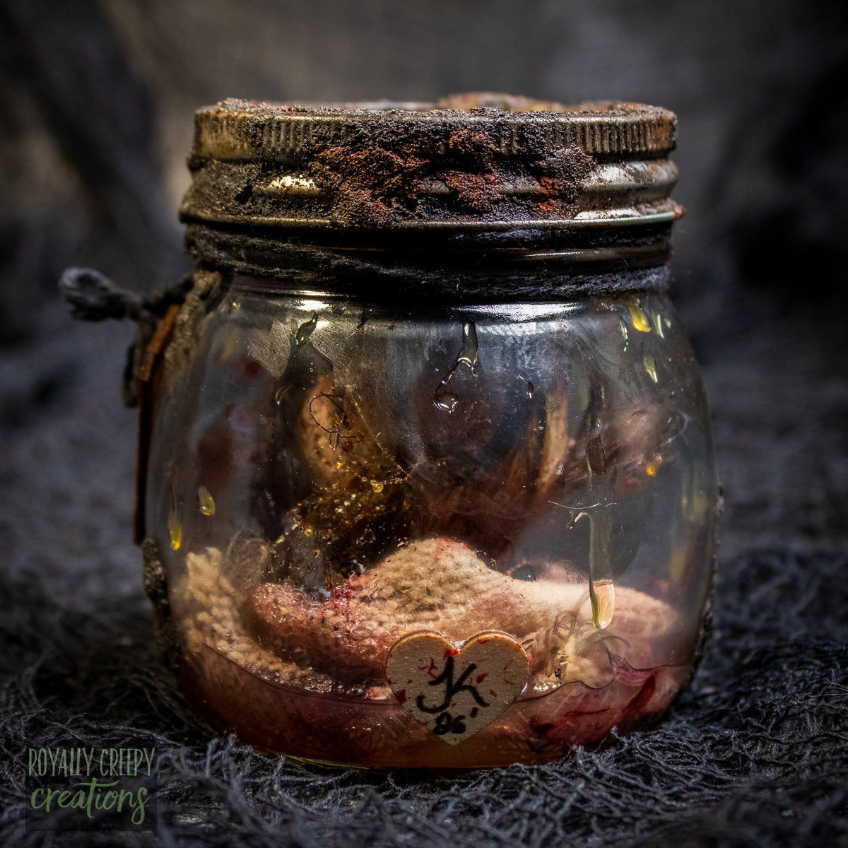 A creepy specimen prop of a severed doll head inside a dirty, rusted glass jar with a dark lid and rope, set against a dark textured background.