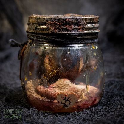 A creepy specimen prop of a severed doll head inside a dirty, rusted glass jar with a dark lid and rope, set against a dark textured background.