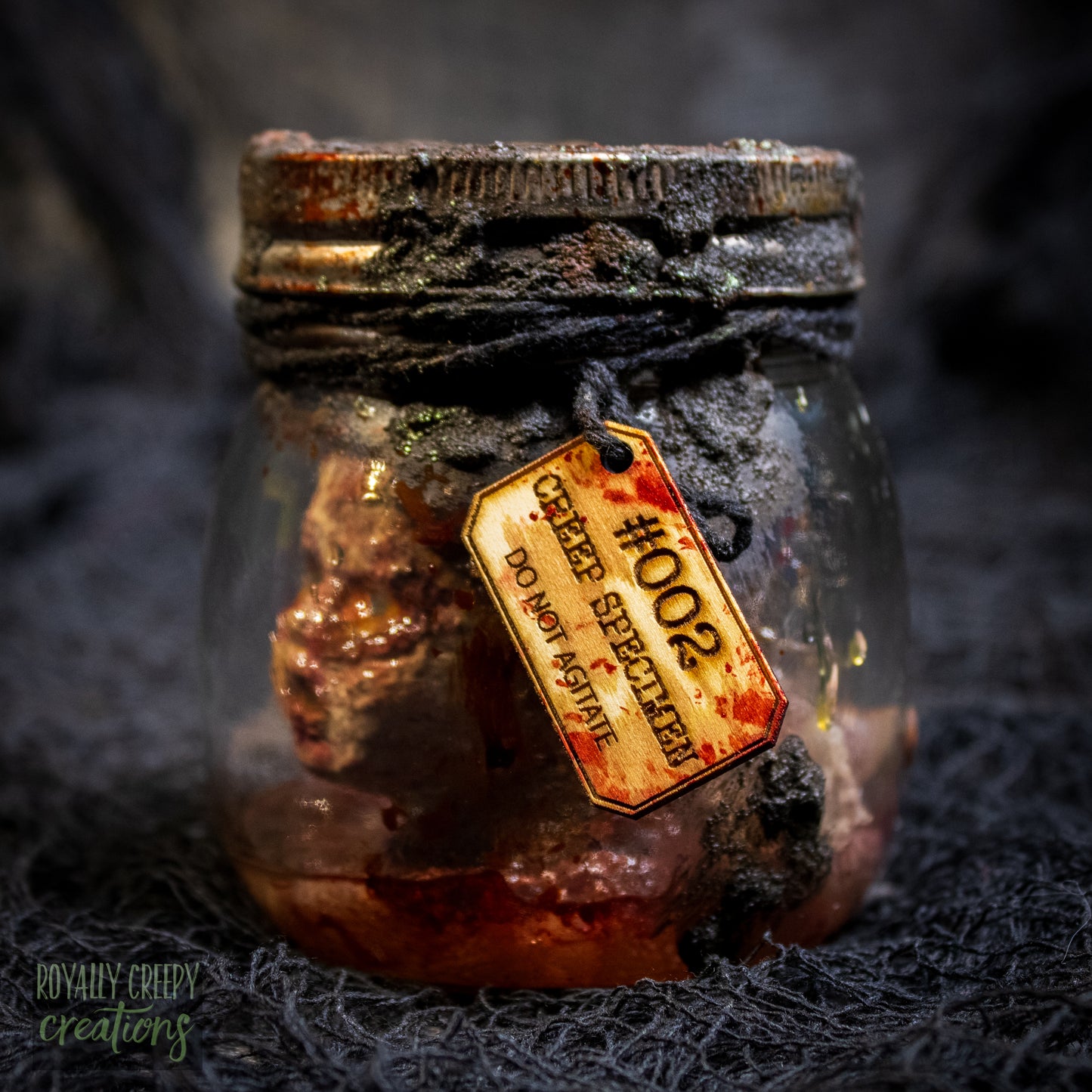 A creepy specimen prop of a severed doll head inside a dirty, rusted glass jar with a dark lid and rope, set against a dark textured background.