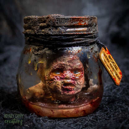 A creepy specimen prop of a severed doll head inside a dirty, rusted glass jar with a dark lid and rope, set against a dark textured background.