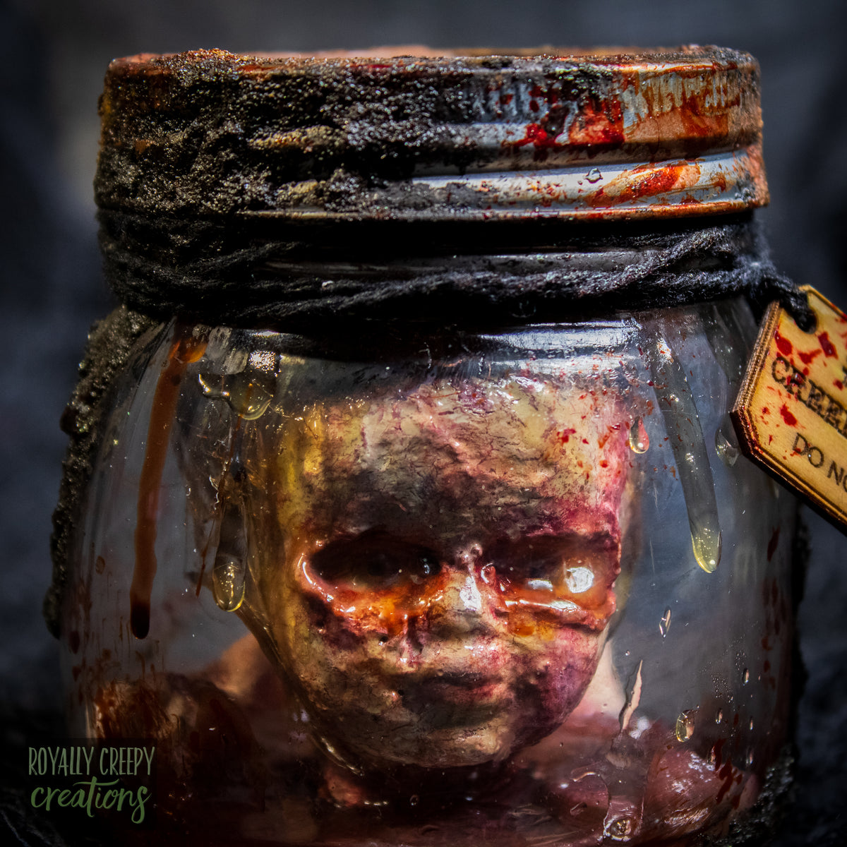 A creepy specimen prop of a severed doll head inside a dirty, rusted glass jar with a dark lid and rope, set against a dark textured background.