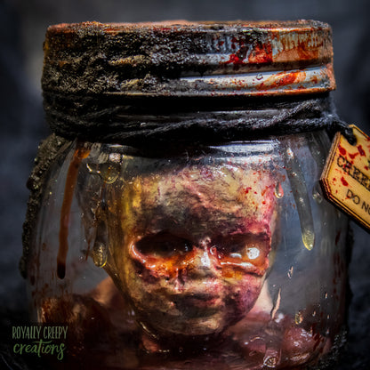 A creepy specimen prop of a severed doll head inside a dirty, rusted glass jar with a dark lid and rope, set against a dark textured background.
