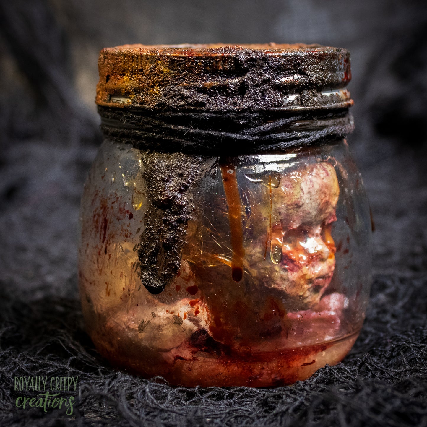 A creepy specimen prop of a severed doll head inside a dirty, rusted glass jar with a dark lid and rope, set against a dark textured background.