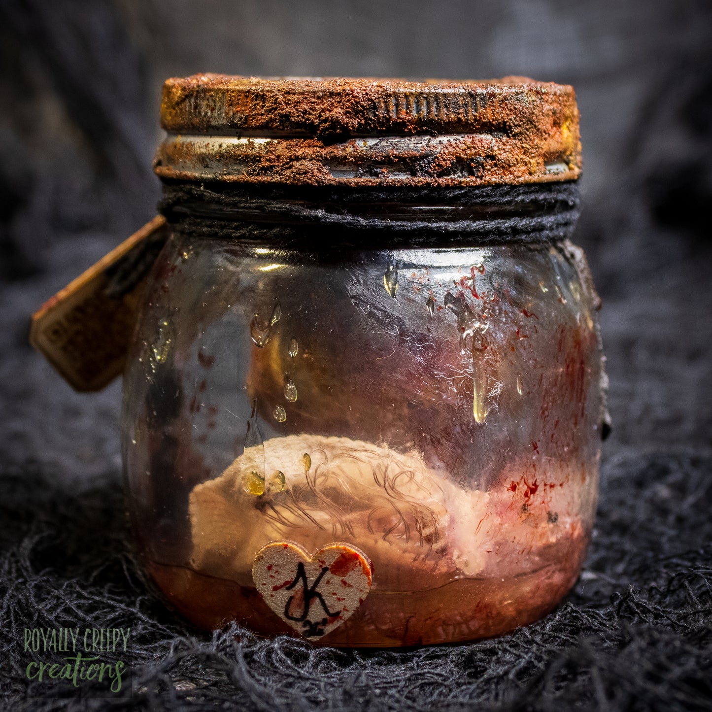 A creepy specimen prop of a severed doll head inside a dirty, rusted glass jar with a dark lid and rope, set against a dark textured background.