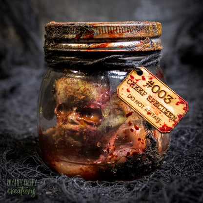 A creepy specimen prop of a severed doll head inside a dirty, rusted glass jar with a dark lid and rope, set against a dark textured background.