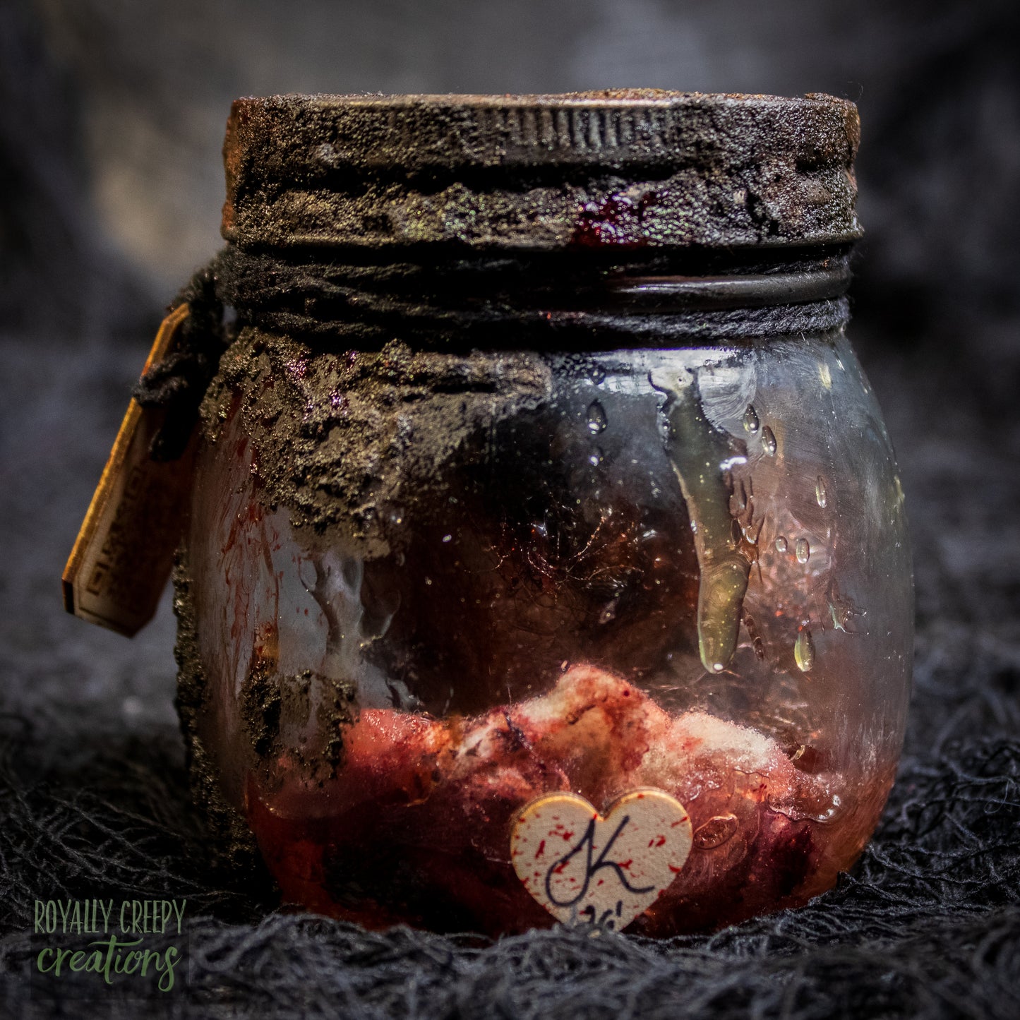 A creepy specimen prop of a severed doll head inside a dirty, rusted glass jar with a dark lid and rope, set against a dark textured background.