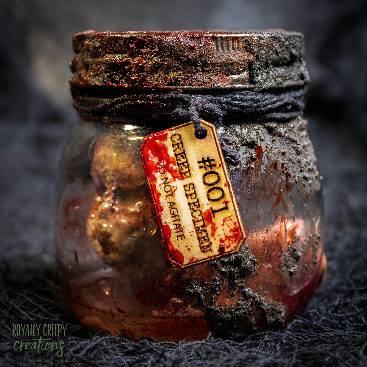 A creepy specimen prop of a severed doll head inside a dirty, rusted glass jar with a dark lid and rope, set against a dark textured background.