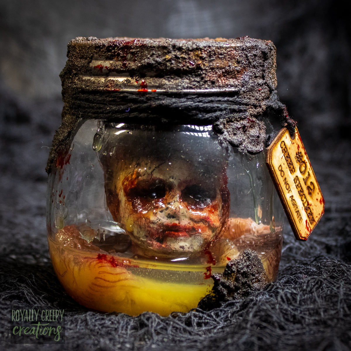 A creepy specimen prop of a severed doll head inside a dirty, rusted glass jar with a dark lid and rope, set against a dark textured background.