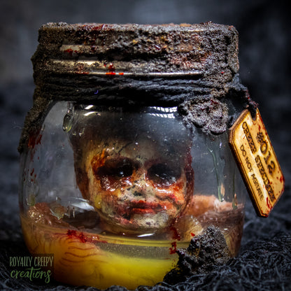 A creepy specimen prop of a severed doll head inside a dirty, rusted glass jar with a dark lid and rope, set against a dark textured background.