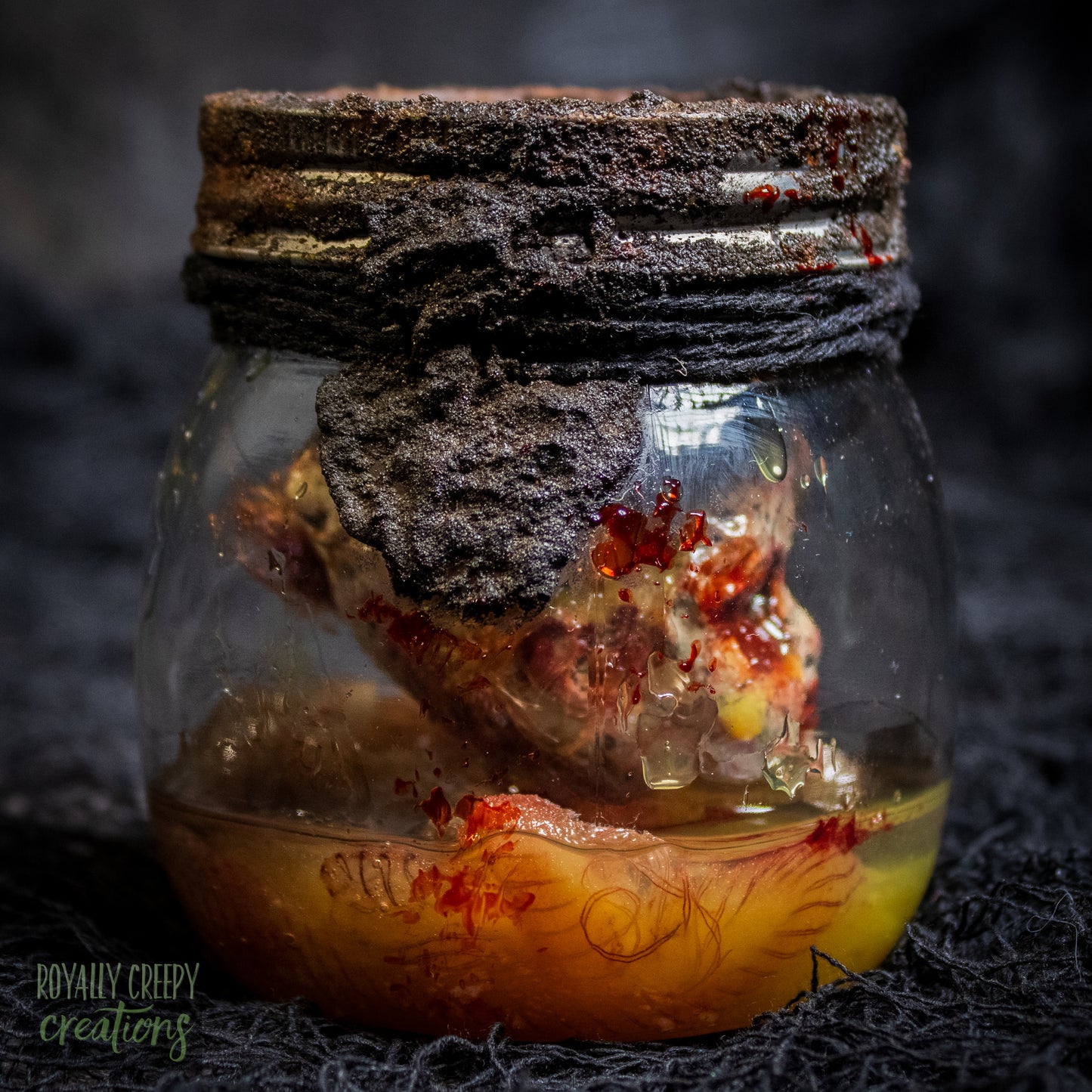 A creepy specimen prop of a severed doll head inside a dirty, rusted glass jar with a dark lid and rope, set against a dark textured background.