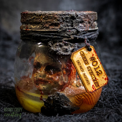 A creepy specimen prop of a severed doll head inside a dirty, rusted glass jar with a dark lid and rope, set against a dark textured background.
