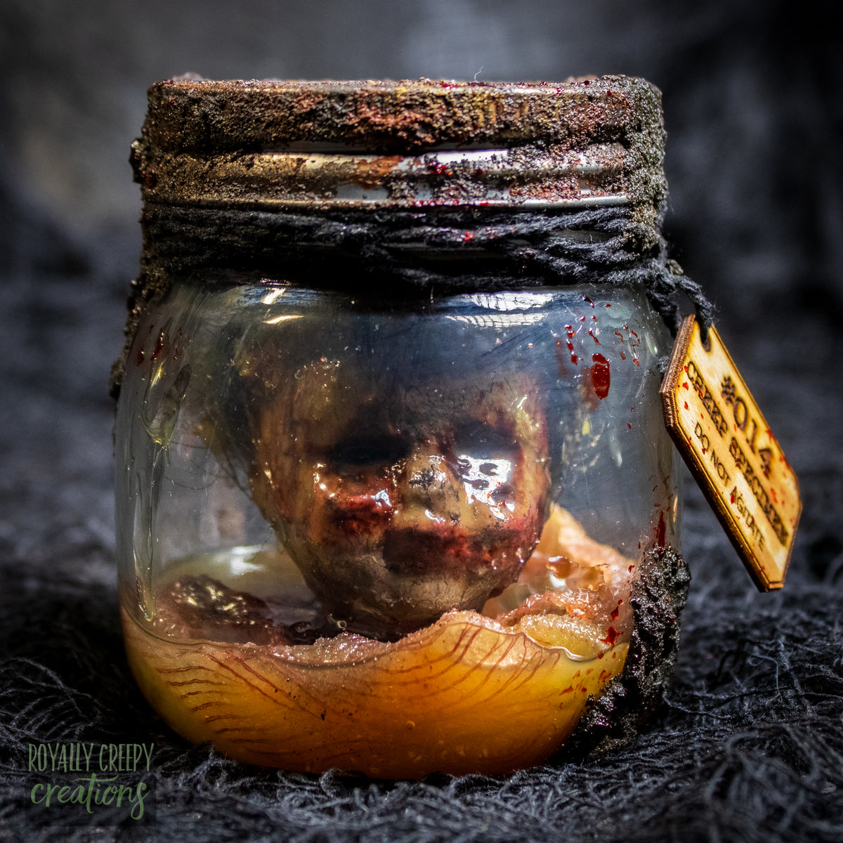 A creepy specimen prop of a severed doll head inside a dirty, rusted glass jar with a dark lid and rope, set against a dark textured background.