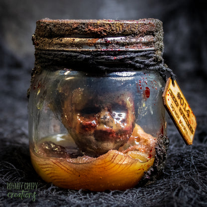 A creepy specimen prop of a severed doll head inside a dirty, rusted glass jar with a dark lid and rope, set against a dark textured background.