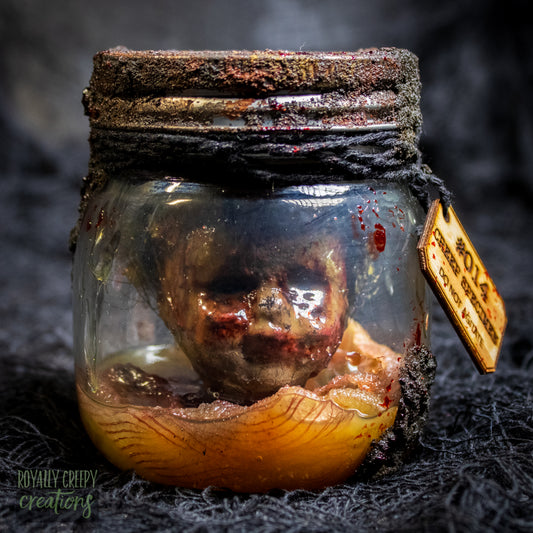 A creepy specimen prop of a severed doll head inside a dirty, rusted glass jar with a dark lid and rope, set against a dark textured background.