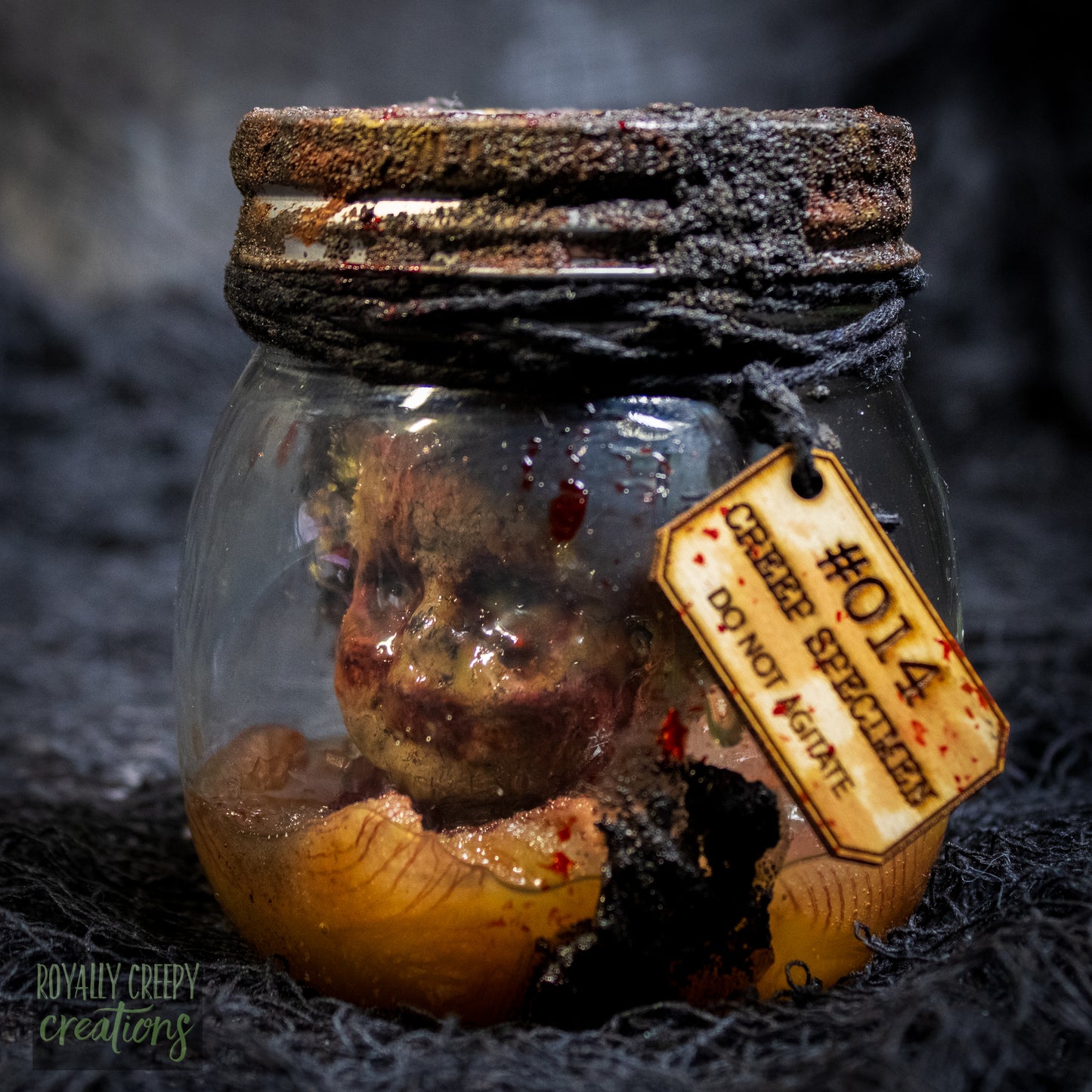 A creepy specimen prop of a severed doll head inside a dirty, rusted glass jar with a dark lid and rope, set against a dark textured background.