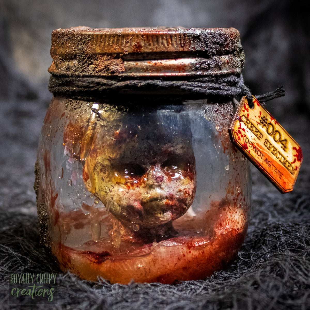 A creepy specimen prop of a severed doll head inside a dirty, rusted glass jar with a dark lid and rope, set against a dark textured background.