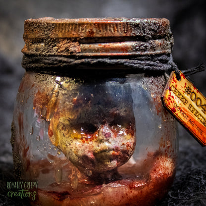 A creepy specimen prop of a severed doll head inside a dirty, rusted glass jar with a dark lid and rope, set against a dark textured background.