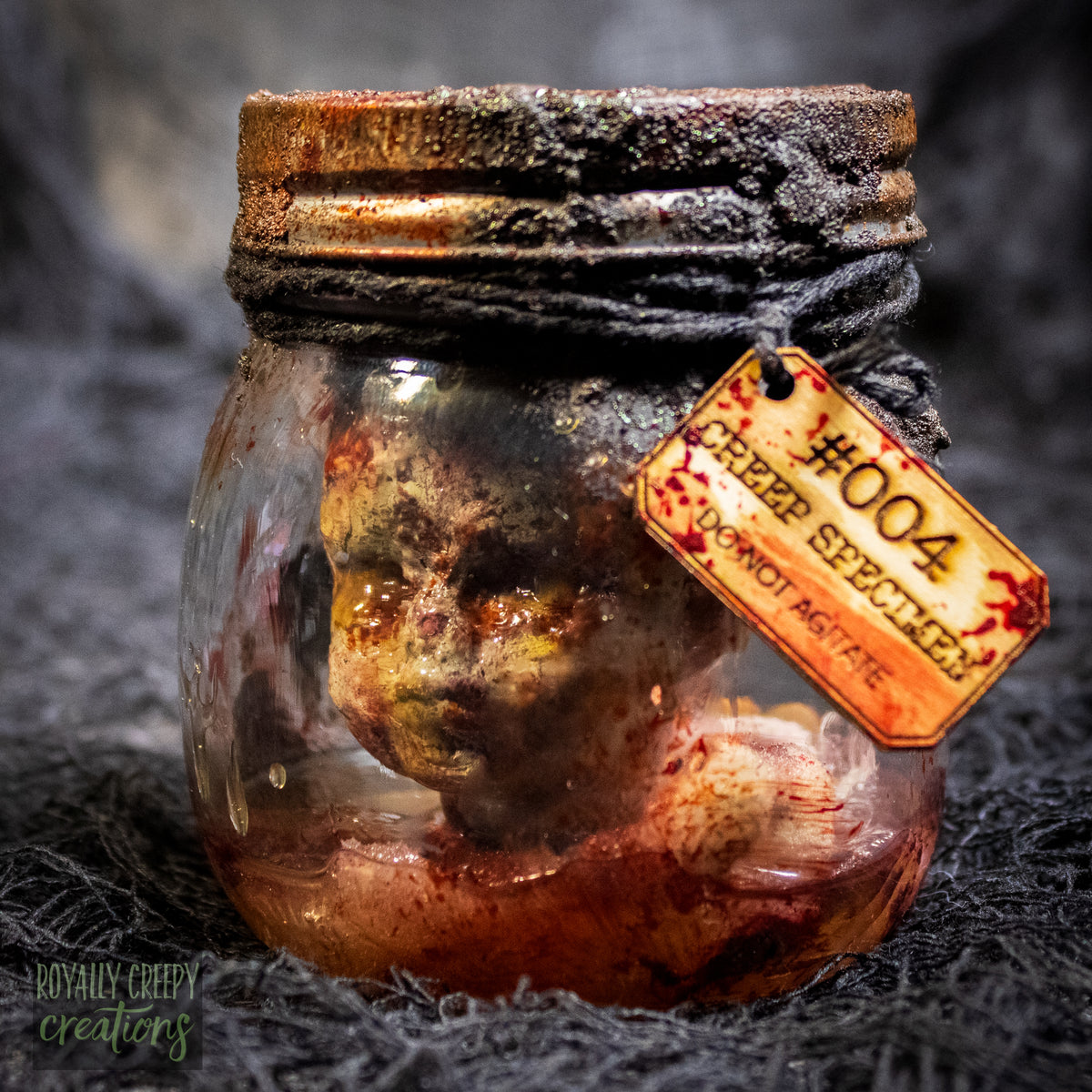 A creepy specimen prop of a severed doll head inside a dirty, rusted glass jar with a dark lid and rope, set against a dark textured background.
