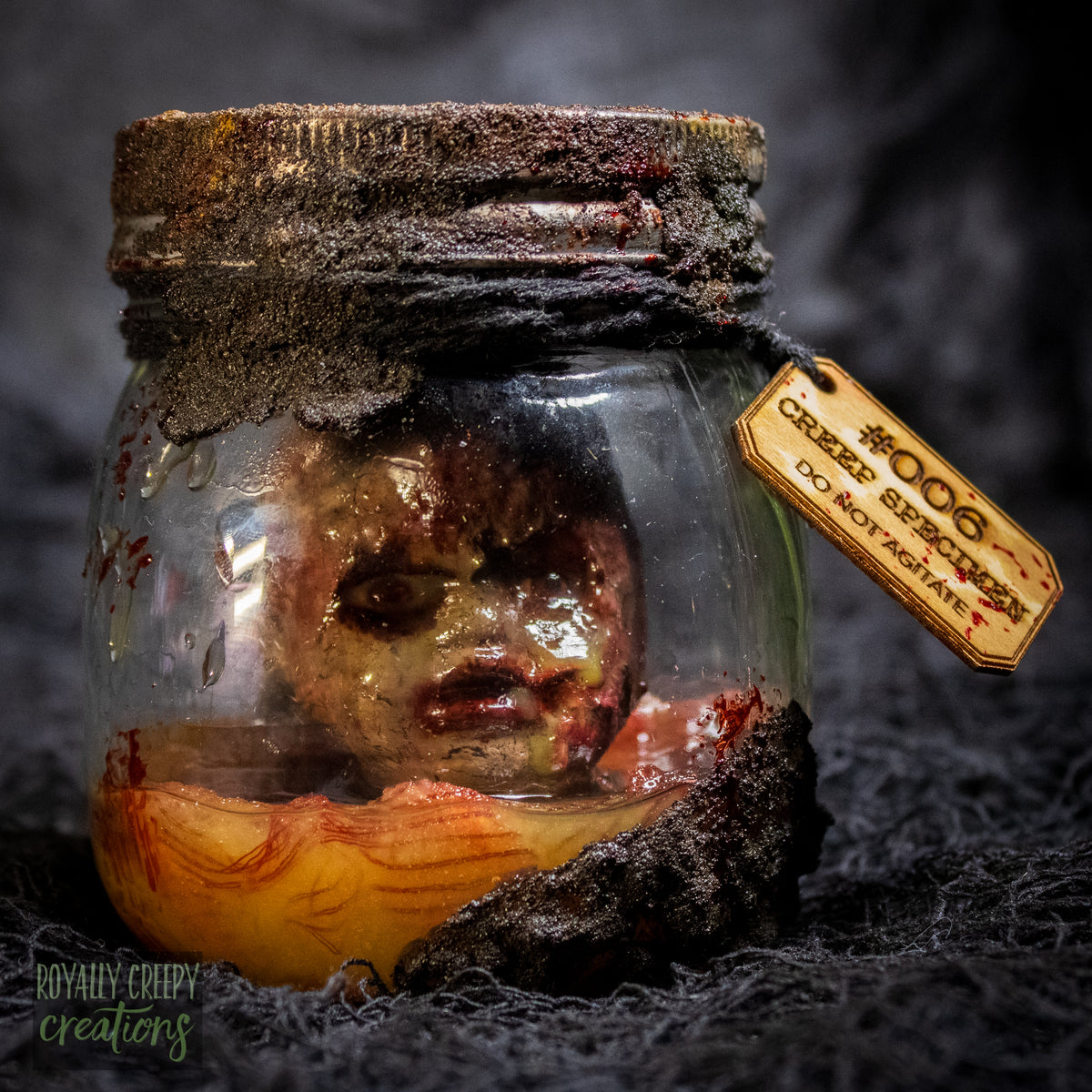 A creepy specimen prop of a severed doll head inside a dirty, rusted glass jar with a dark lid and rope, set against a dark textured background.