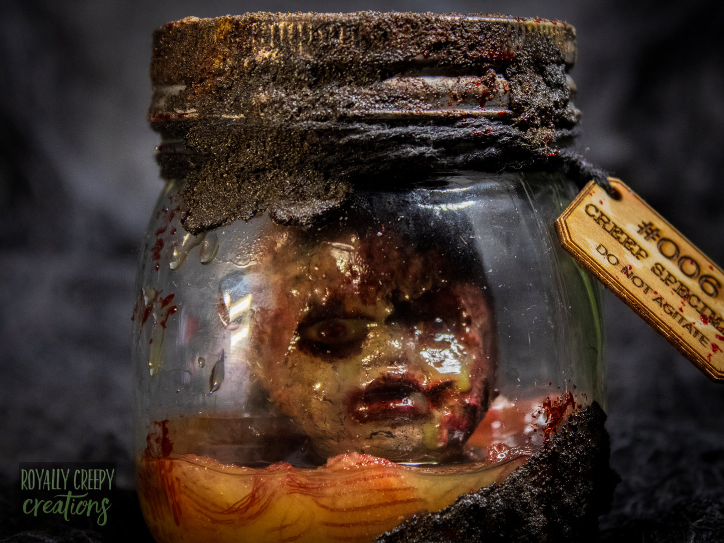 A creepy specimen prop of a severed doll head inside a dirty, rusted glass jar with a dark lid and rope, set against a dark textured background.