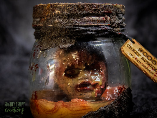 A creepy specimen prop of a severed doll head inside a dirty, rusted glass jar with a dark lid and rope, set against a dark textured background.