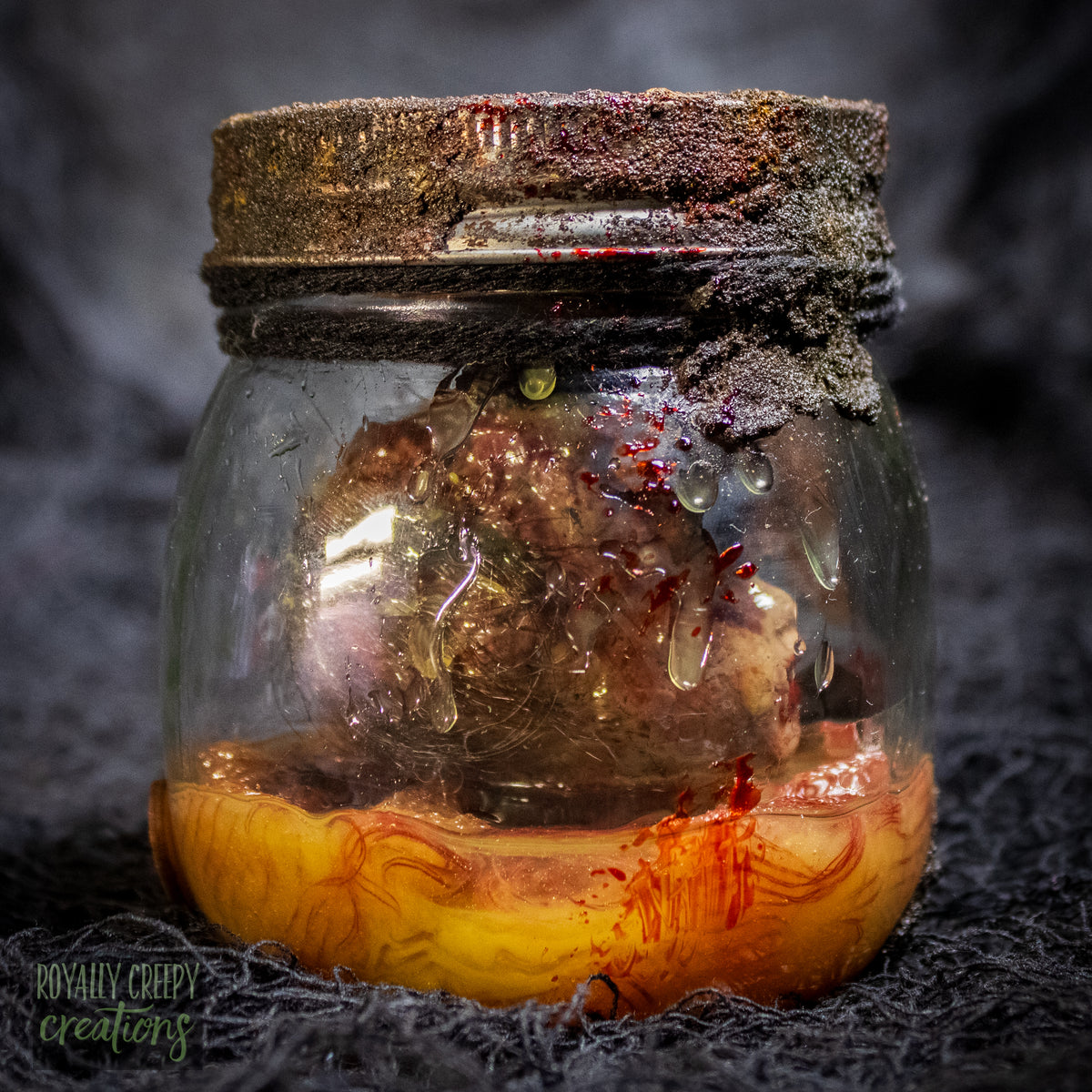 A creepy specimen prop of a severed doll head inside a dirty, rusted glass jar with a dark lid and rope, set against a dark textured background.