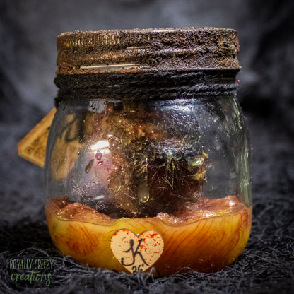 A creepy specimen prop of a severed doll head inside a dirty, rusted glass jar with a dark lid and rope, set against a dark textured background.