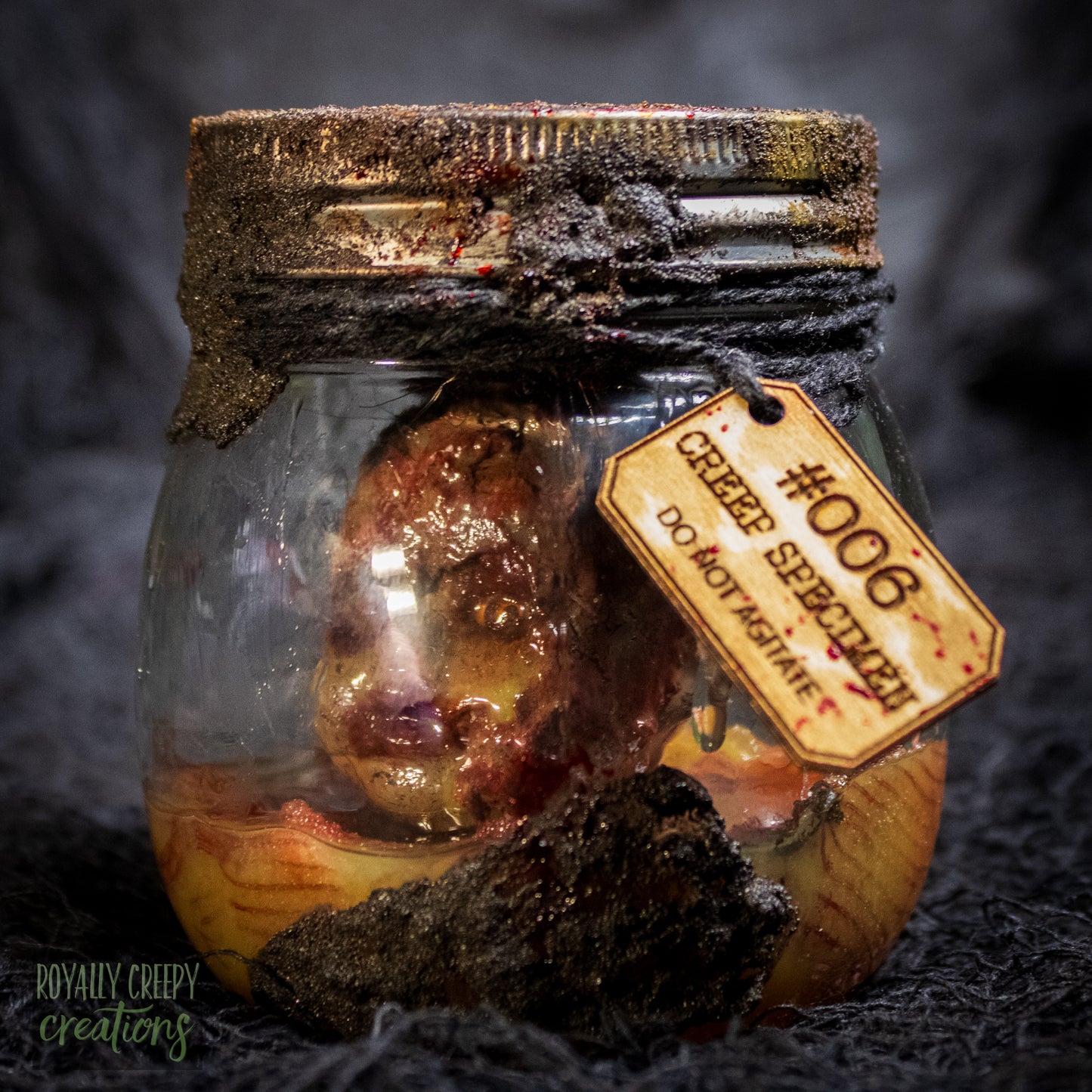 A creepy specimen prop of a severed doll head inside a dirty, rusted glass jar with a dark lid and rope, set against a dark textured background.