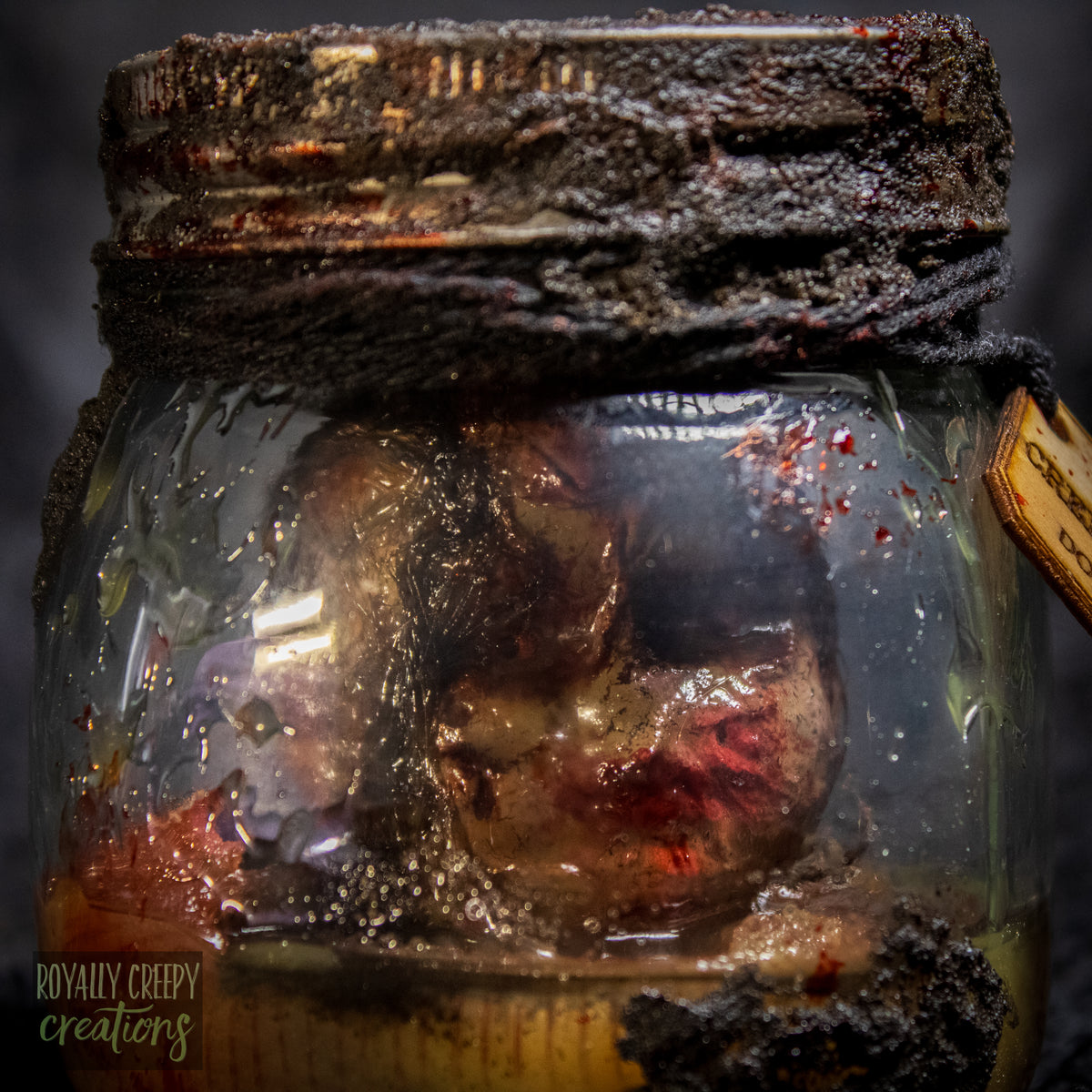 A creepy specimen prop of a severed doll head inside a dirty, rusted glass jar with a dark lid and rope, set against a dark textured background.