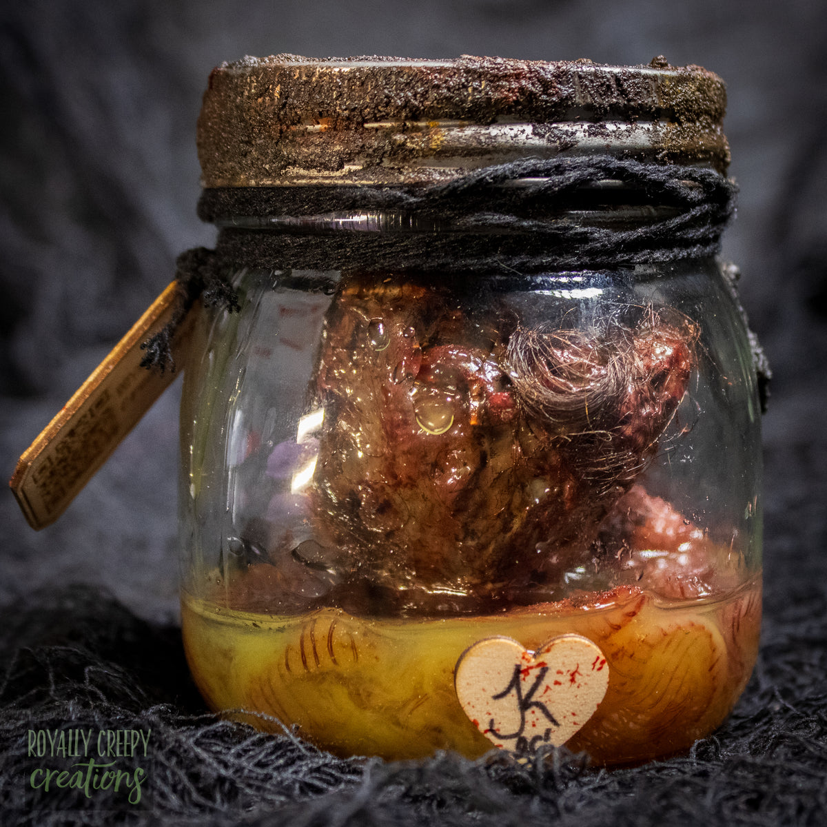 A creepy specimen prop of a severed doll head inside a dirty, rusted glass jar with a dark lid and rope, set against a dark textured background.