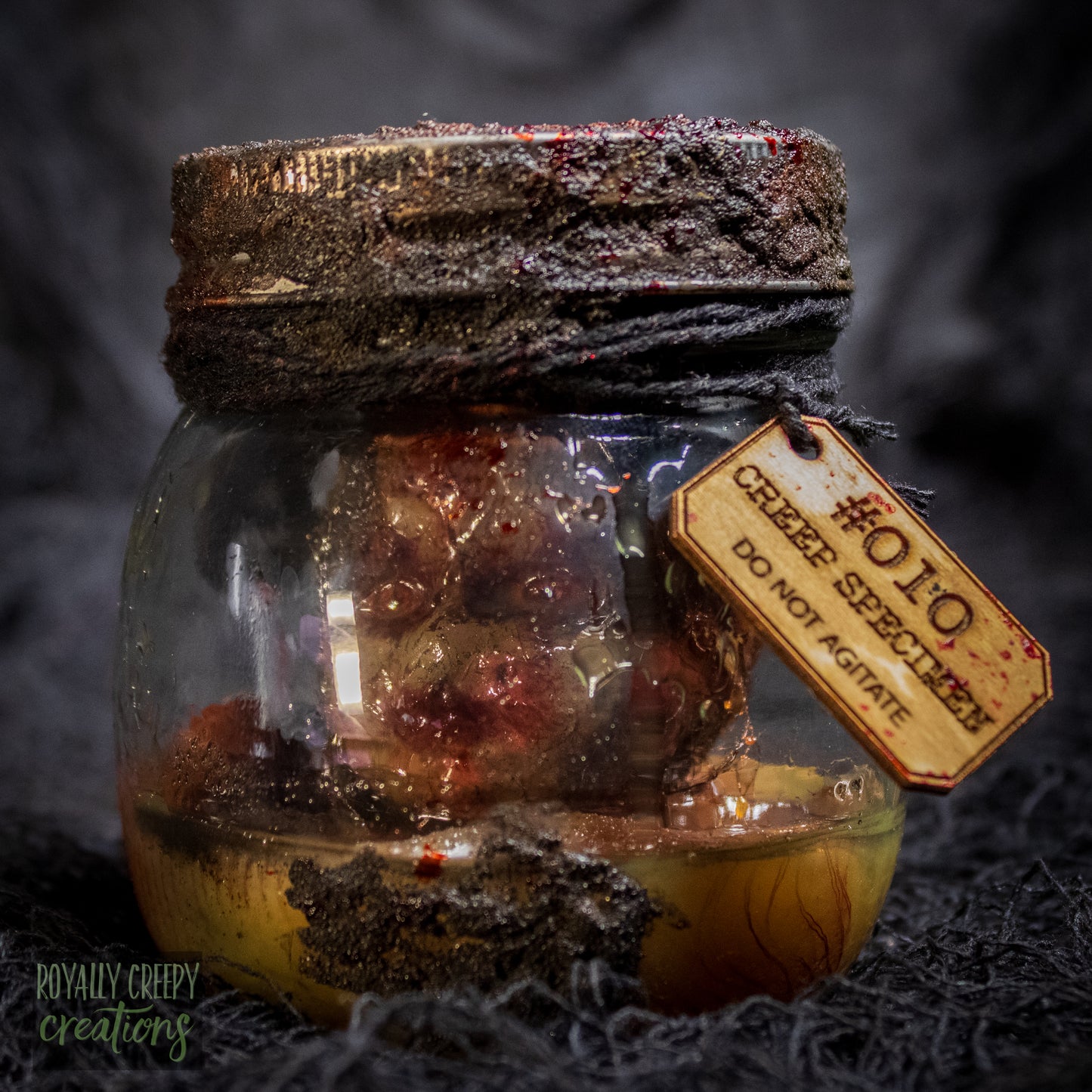 A creepy specimen prop of a severed doll head inside a dirty, rusted glass jar with a dark lid and rope, set against a dark textured background.