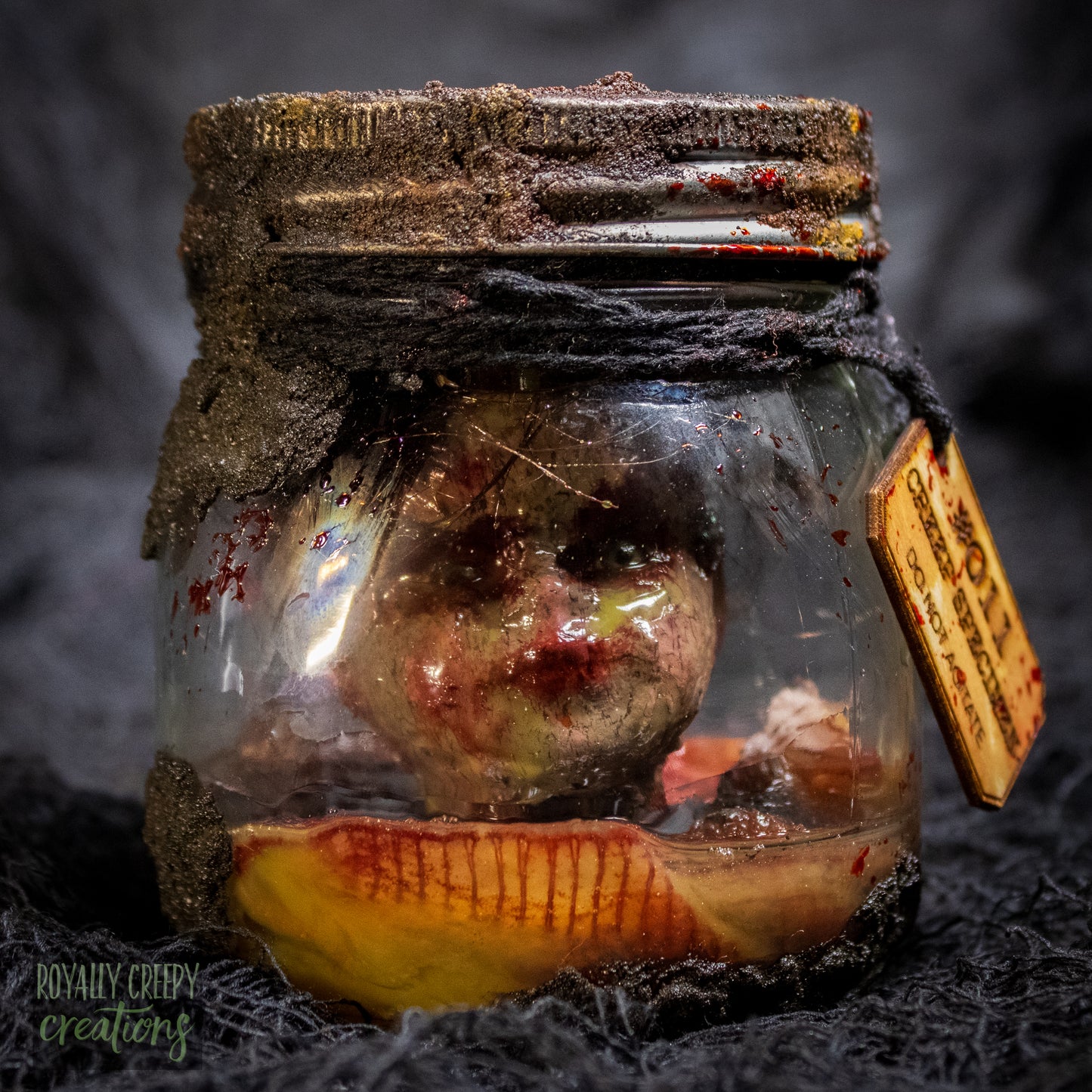 A creepy specimen prop of a severed doll head inside a dirty, rusted glass jar with a dark lid and rope, set against a dark textured background.