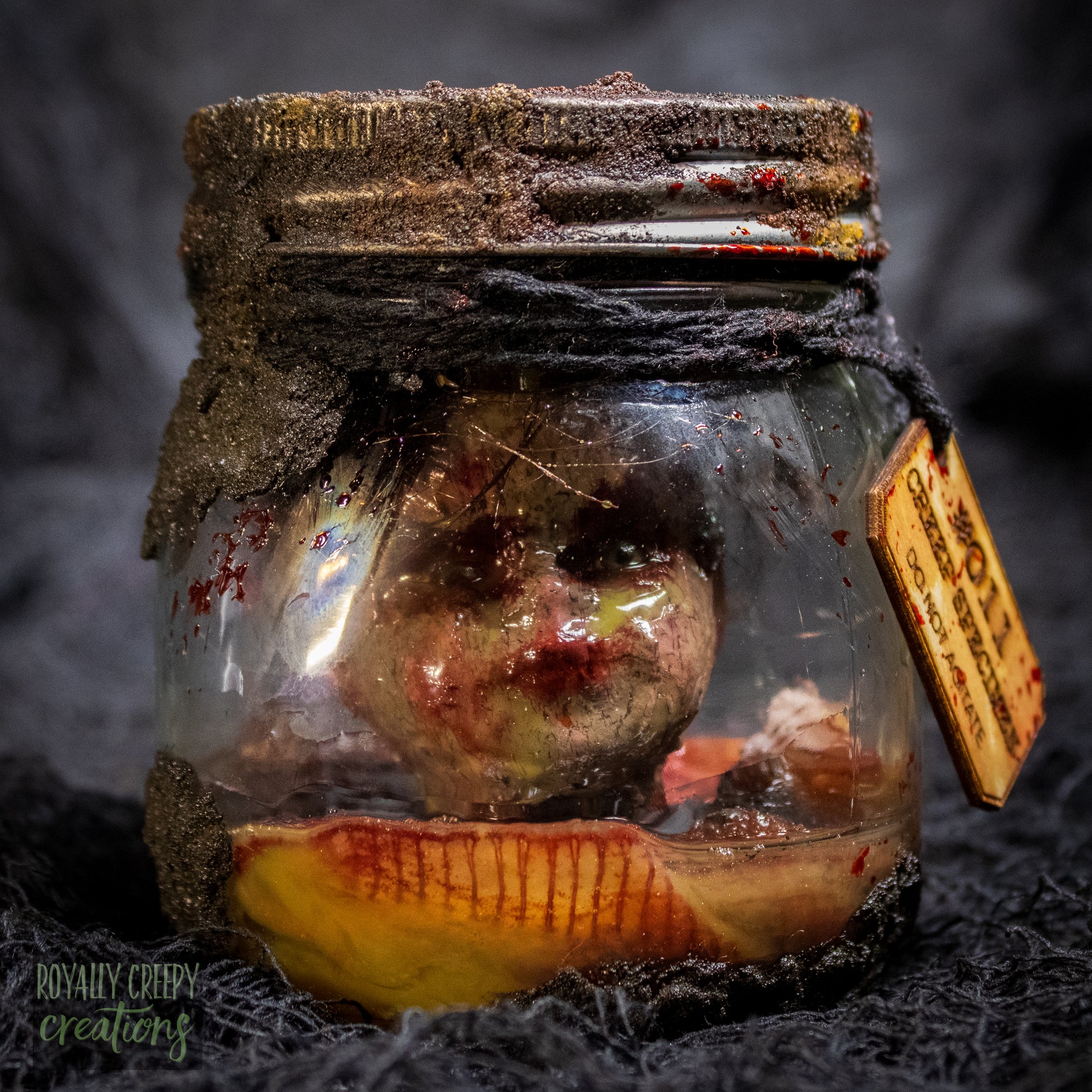 A creepy specimen prop of a severed doll head inside a dirty, rusted glass jar with a dark lid and rope, set against a dark textured background.