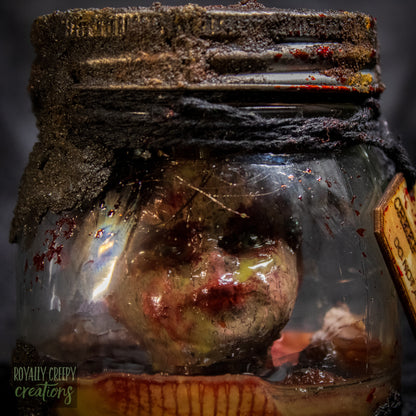 A creepy specimen prop of a severed doll head inside a dirty, rusted glass jar with a dark lid and rope, set against a dark textured background.