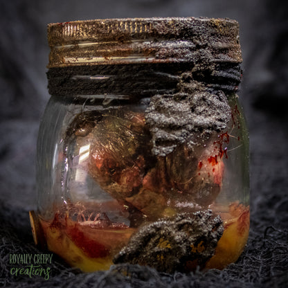 A creepy specimen prop of a severed doll head inside a dirty, rusted glass jar with a dark lid and rope, set against a dark textured background.