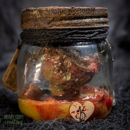 A creepy specimen prop of a severed doll head inside a dirty, rusted glass jar with a dark lid and rope, set against a dark textured background.